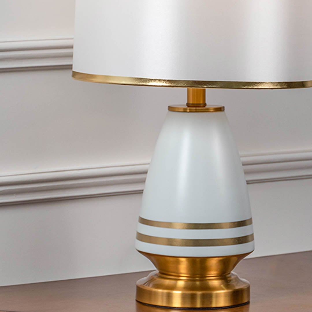 Closeup of white ceramic table lamp with golden base striped detailing and fabric drum shade for modern decor.