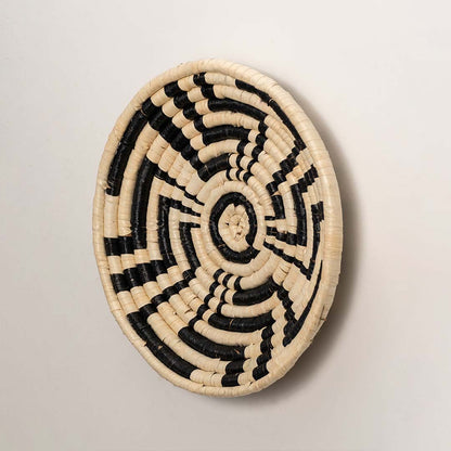 A side angle view of the natural and black rattan wall decor plate showing its raised woven rim and textured artisan finish for warm rustic home wall styling.