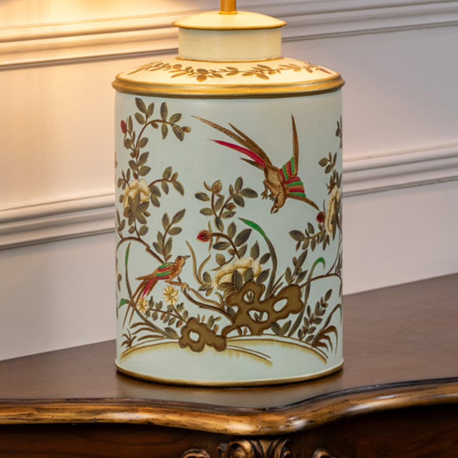 Table lamp closeup showing hand painted bird and floral design on ceramic base with soft green background and gold rim.