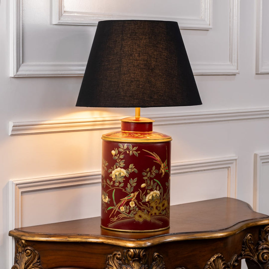 Red ceramic table lamp with floral bird artwork and black fabric shade placed on carved wooden console adding warm classic lighting.