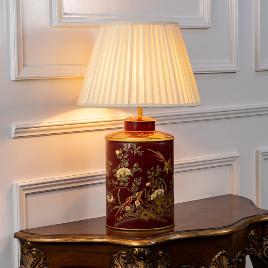 Light on white background shows red ceramic table lamp glowing warmly through black fabric shade highlighting floral bird artwork.