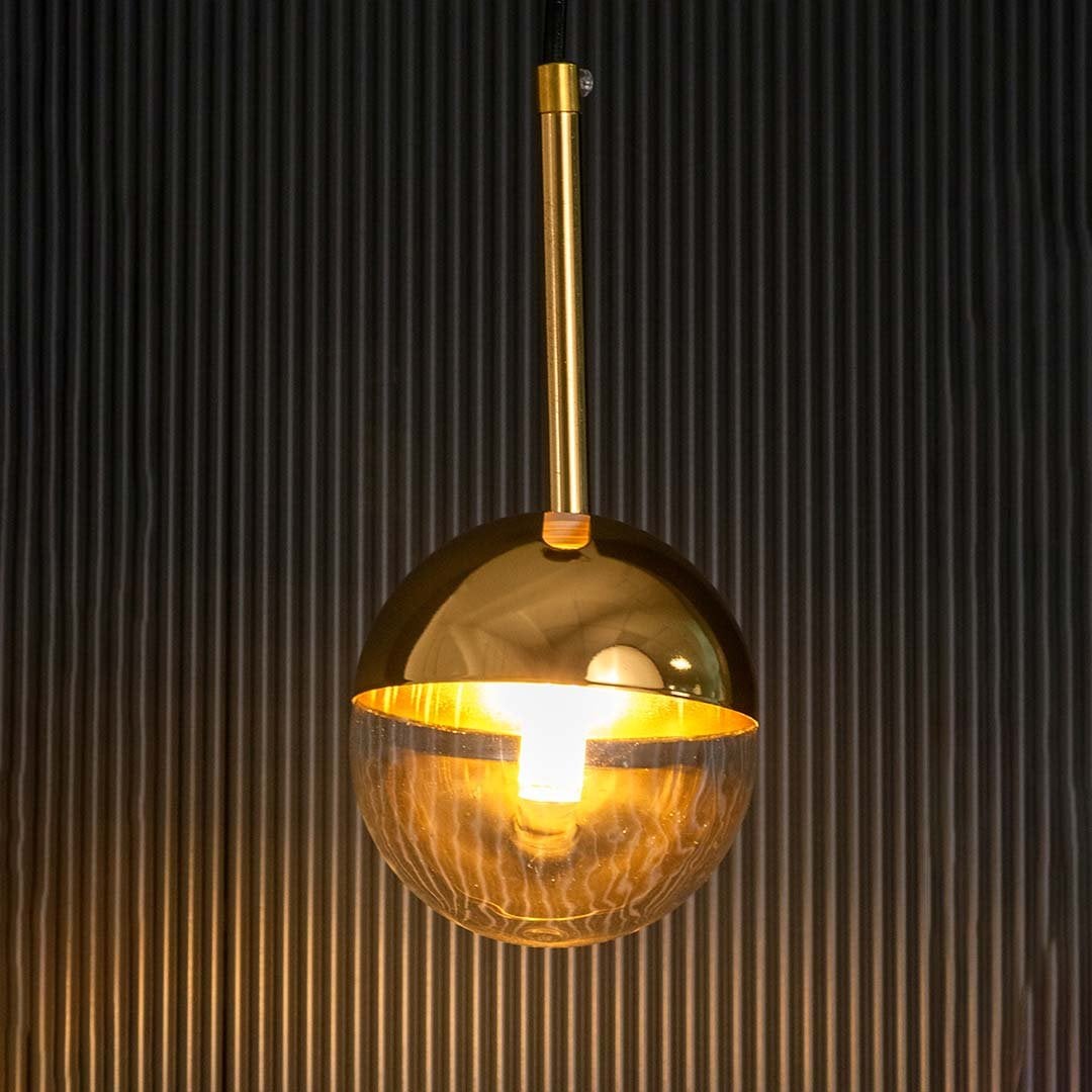 A gold and glass spherical pendant light glowing warmly with the light on against a dark black background enhancing the reflective golden surface and clear globe.