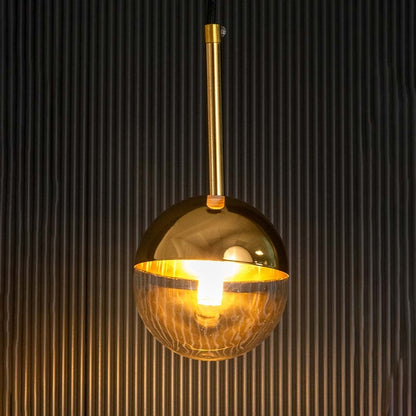 A gold and glass spherical pendant light glowing warmly with the light on against a dark black background enhancing the reflective golden surface and clear globe.