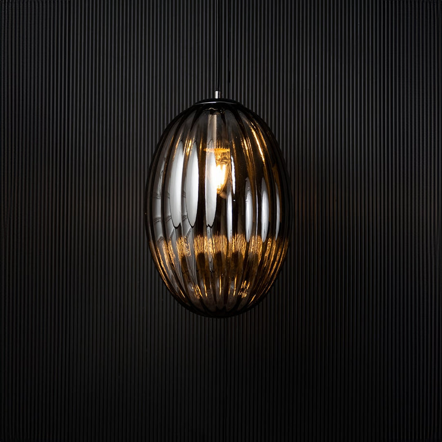 A ribbed oval glass pendant light shown with the light on against a dark background creating a soft glowing effect that enhances the elegant vertical contours.