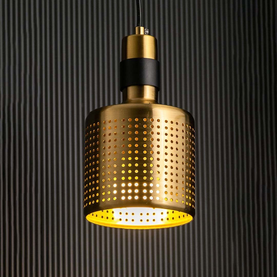 A gold perforated pendant light with lights on casting a patterned warm glow on a dark backdrop showcasing its cylindrical metal design.