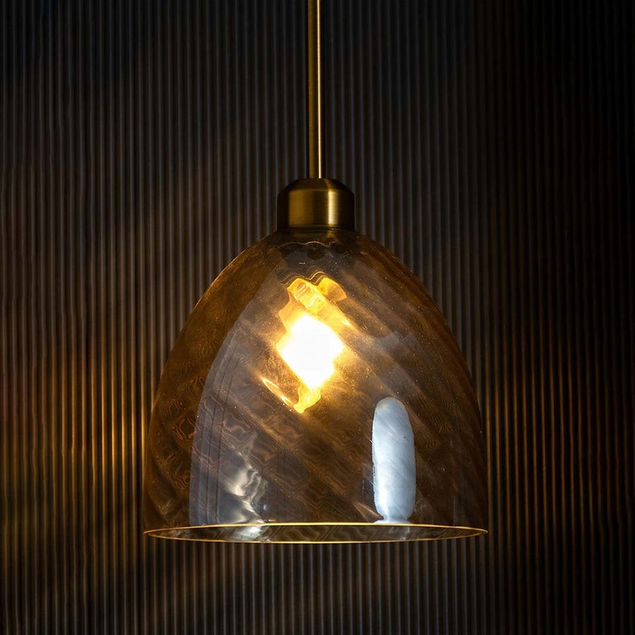 A gold and smoked glass pendant light with lights on casting a warm glow on a black backdrop showcasing its textured smoky dome shade.