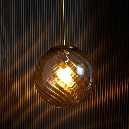 A gold and amber pendant light with lights on casting a warm amber glow on a black backdrop showcasing its round textured glass shade.
