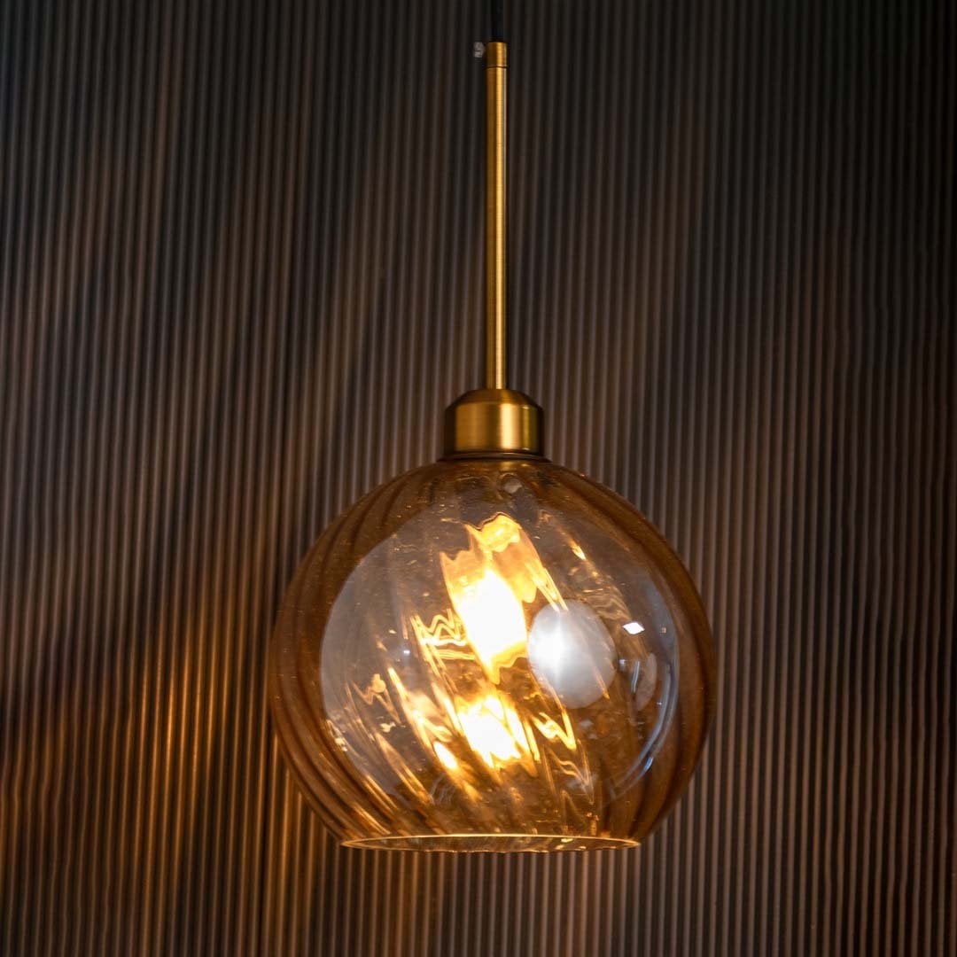 A smoked glass pendant with gold metal rod and bulb on casting warm ambient light against a deep black backdrop highlighting its modern globe design.