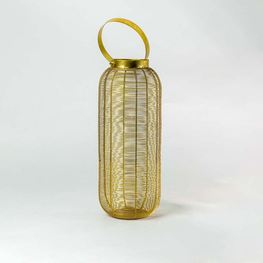 Gold metal wire mesh lantern with candle light glowing on outdoor garden patio with plants and fairy lights in background.