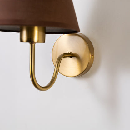 Closeup wall light in gold metal finish with curved arm and fabric shade for bedroom or living decor lighting white background.