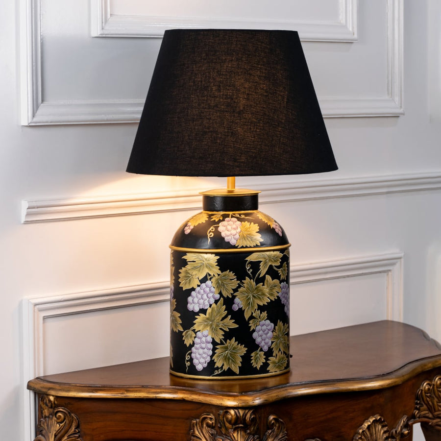 Table lamp with black fabric shade and black ceramic base featuring gold botanical detailing displayed on a wooden table against a white wall background.