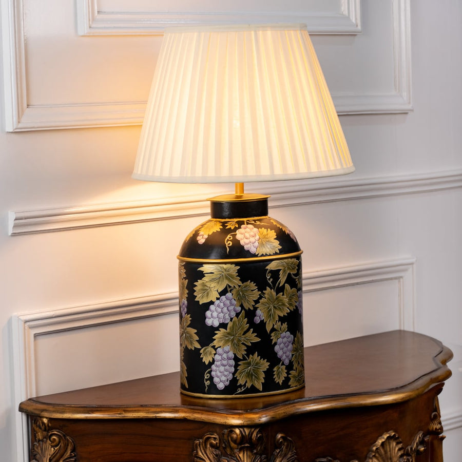 Table lamp with white pleated fabric shade and black ceramic base highlighted with gold floral detailing displayed on a wooden table against a white background.