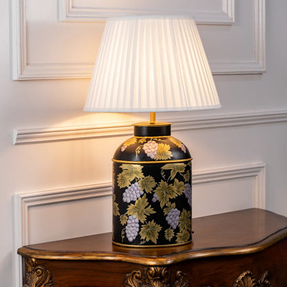 Table lamp with white pleated fabric shade and black ceramic base highlighted with gold floral artwork styled on a wooden table against a white background.