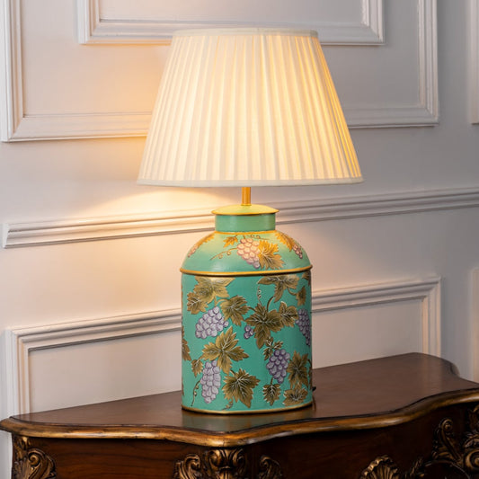 Palais mint ceramic lamp with gold grape design and cream pleated shade glowing warmly on white background highlighting fine detailing.
