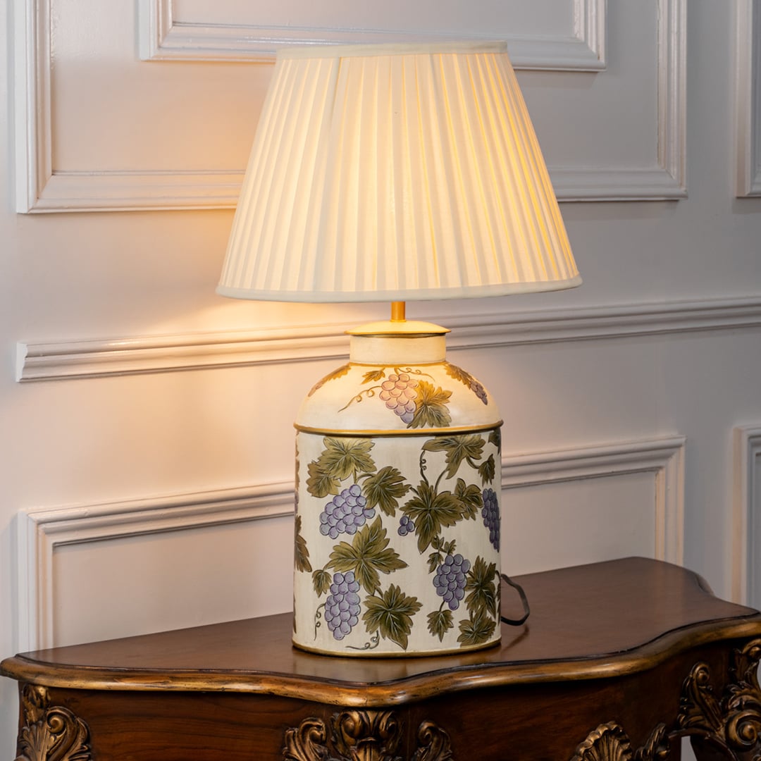 Table lamp on white background highlighting a cream ceramic base decorated with green botanical designs styled with a pleated fabric lampshade.