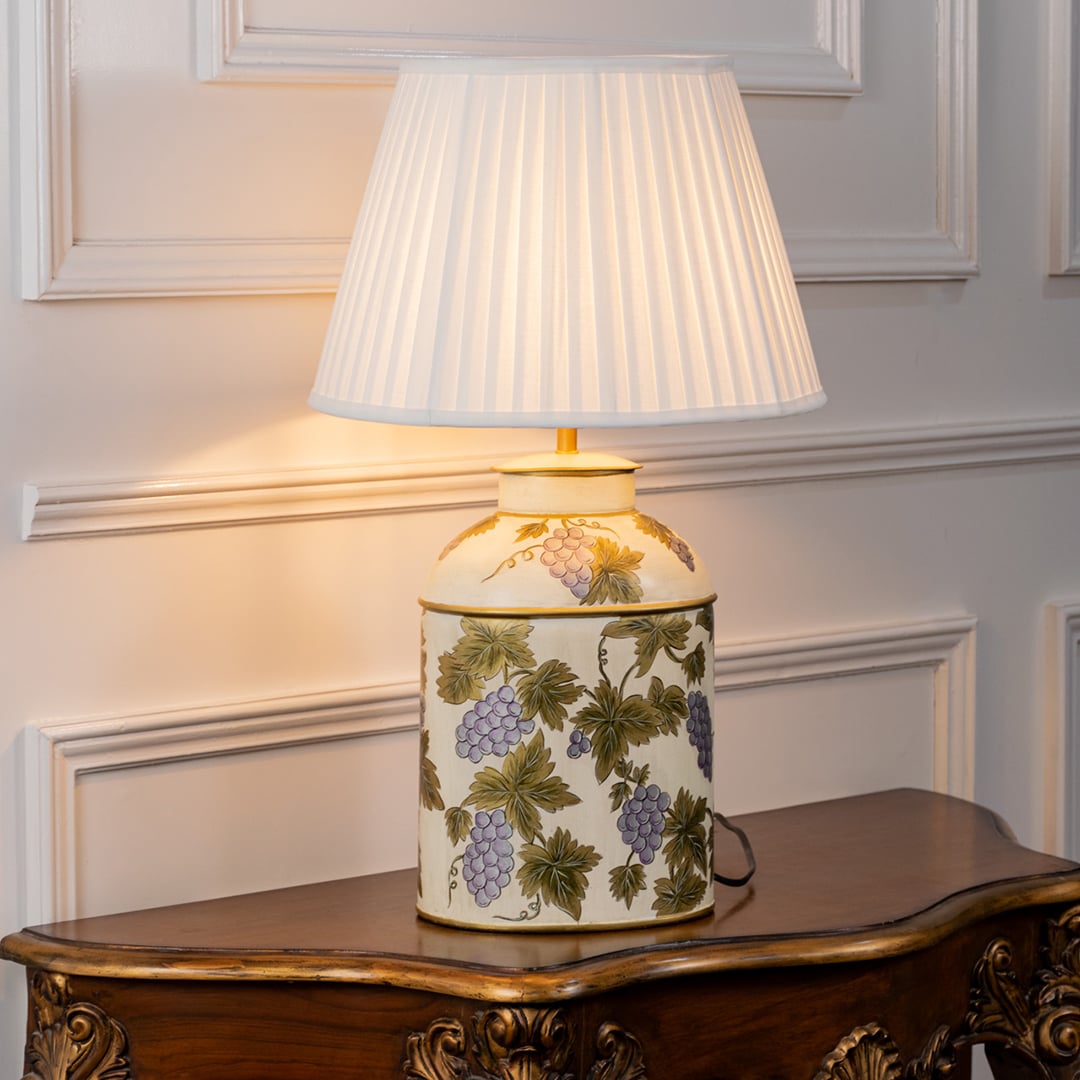 Table lamp with a white pleated shade and cream ceramic base highlighted by green botanical designs placed on a wooden table.