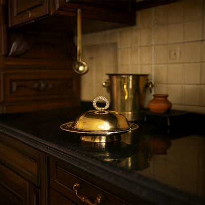 Luxurious gold brass dome serving dish with ring handle placed on a dark kitchen counter next to brass barware for elegant home decor.