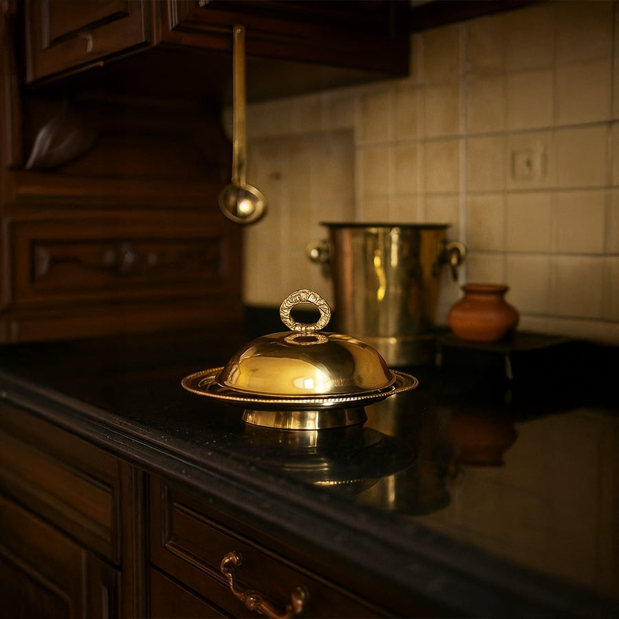 Luxurious gold brass dome serving dish with ring handle placed on a dark kitchen counter next to brass barware for elegant home decor.