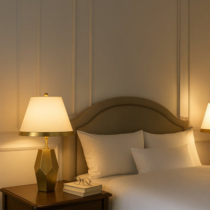 Gold geometric table lamp with a warm white fabric shade placed on a wooden bedside table creating soft ambient lighting in a cozy elegant bedroom setting.