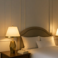 Gold geometric table lamp with a warm white fabric shade placed on a wooden bedside table creating soft ambient lighting in a cozy elegant bedroom setting.
