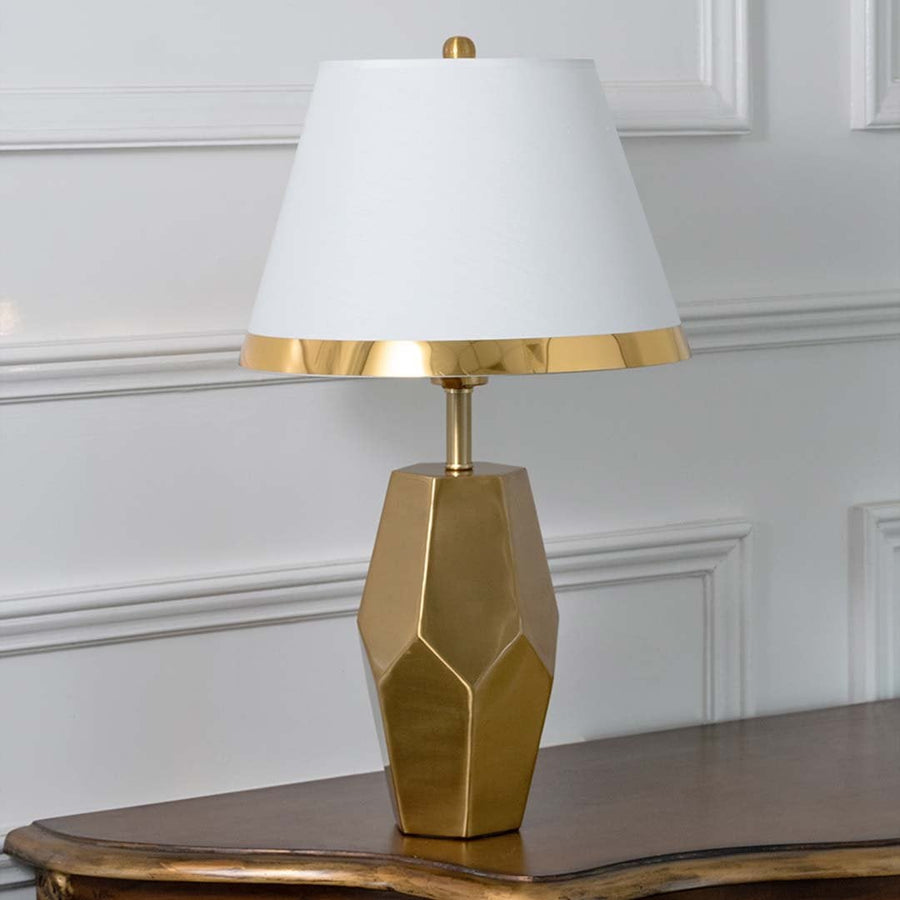 Gold faceted table lamp with white tapered shade and modern sculpted base displayed on a white background perfect for elegant interior styling.