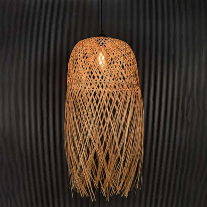 Tall rattan pendant light with bamboo fringe shade turned on glowing softly against plain black background.