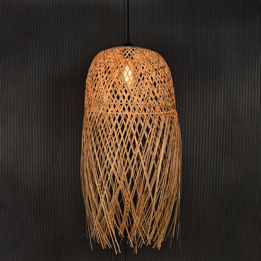 Tall rattan pendant light with bamboo fringe shade turned on glowing softly against plain black background.