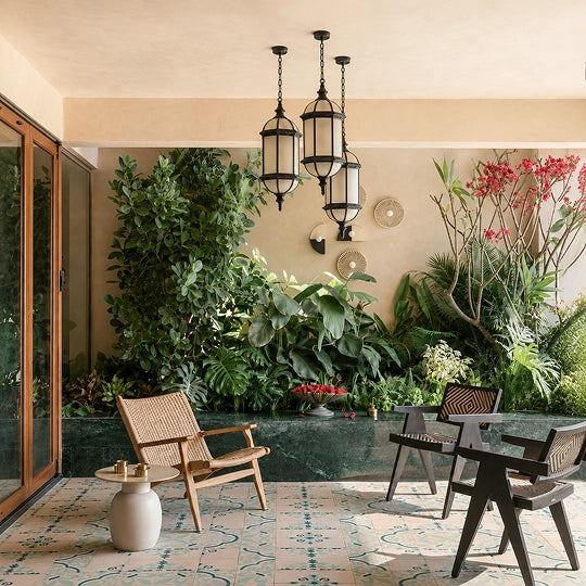 A styled outdoor patio seating area with three modern accent chairs, a small round side table, and three black hanging lanterns, set against a lush green living wall and terracotta-patterned tile flooring.