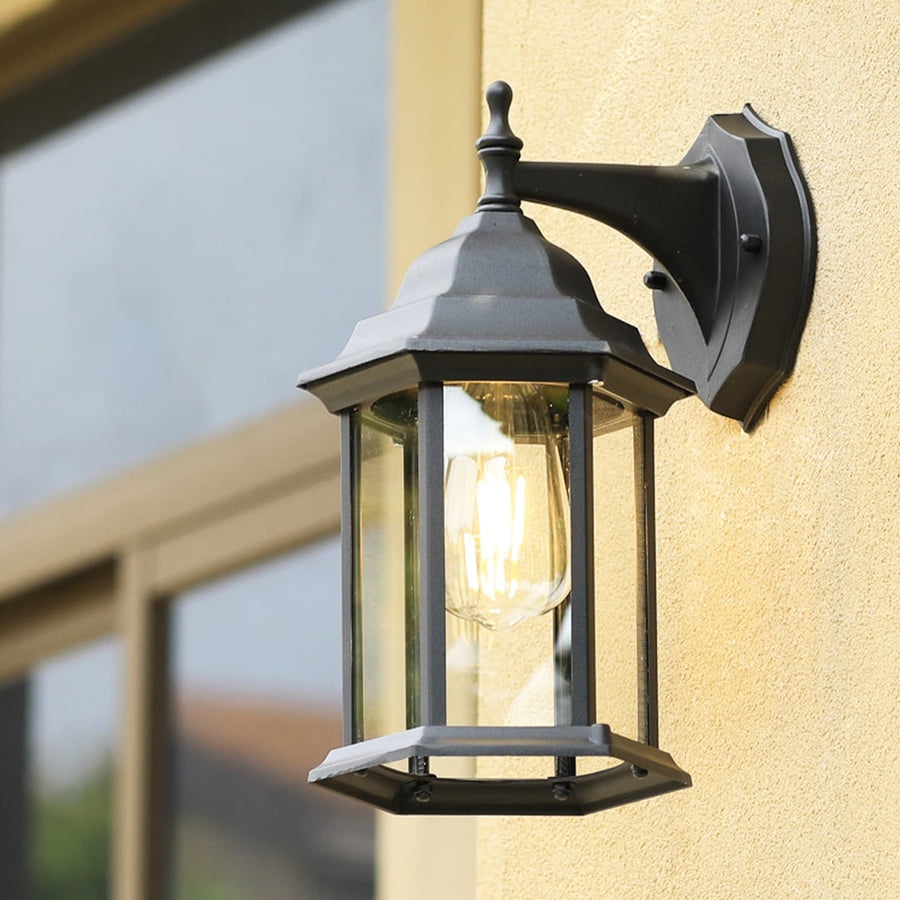 A decorative outdoor wall lantern with the light on mounted near a doorway casting a soft ambient glow on the wall and enhancing the natural greenery around the entrance.