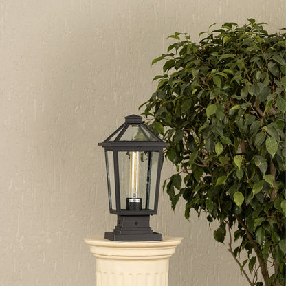 Black outdoor pillar light with clear glass panels placed by a large green plant Lifestyle image patio decor.