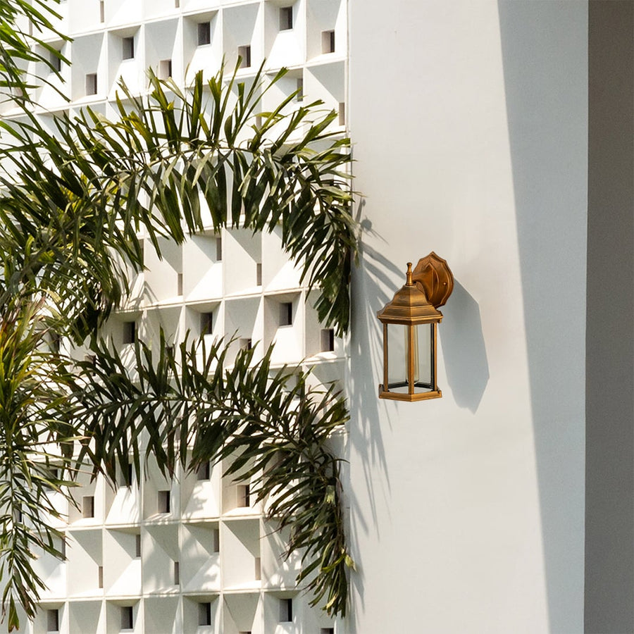 Antique brass outdoor wall lantern lifestyle image with clear glass panels mounted on white villa wall with palm trees.