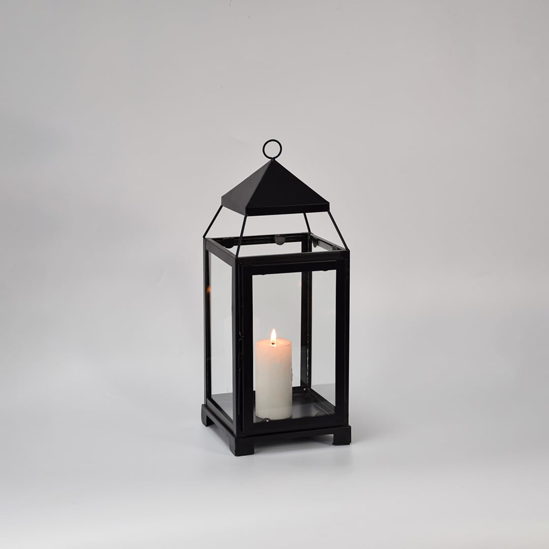 Black metal glass lantern with candle lit shown on white background highlighting warm glow classic frame design and home decor use.