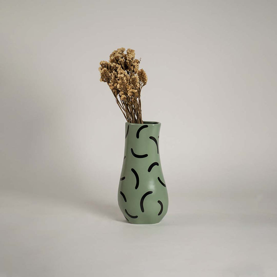 Tall green ceramic vase with black curved patterns holding dried flowers placed on a wooden chest to enhance a cozy vintage themed room.