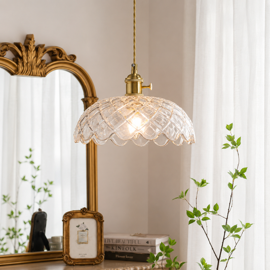 A vintage-style pendant light with a clear scalloped glass shade and a brass-toned ceiling mount, hanging in a softly lit room with a gold mirror, white curtains, and green plant stems.