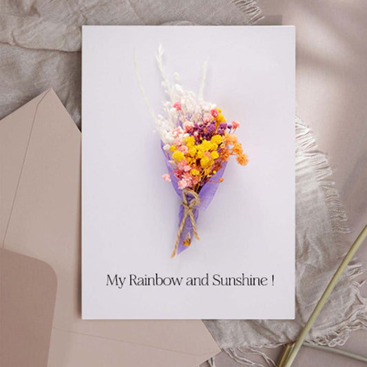 A handmade greeting card featuring a colorful mini dried flower bouquet resting on lace fabric near a window backdrop.