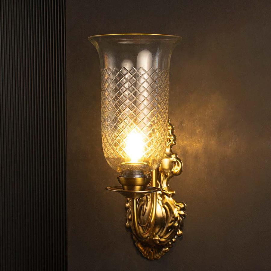 Glowing diamond cut glass wall light with ornate brass base turned on suspended against a dark black background.