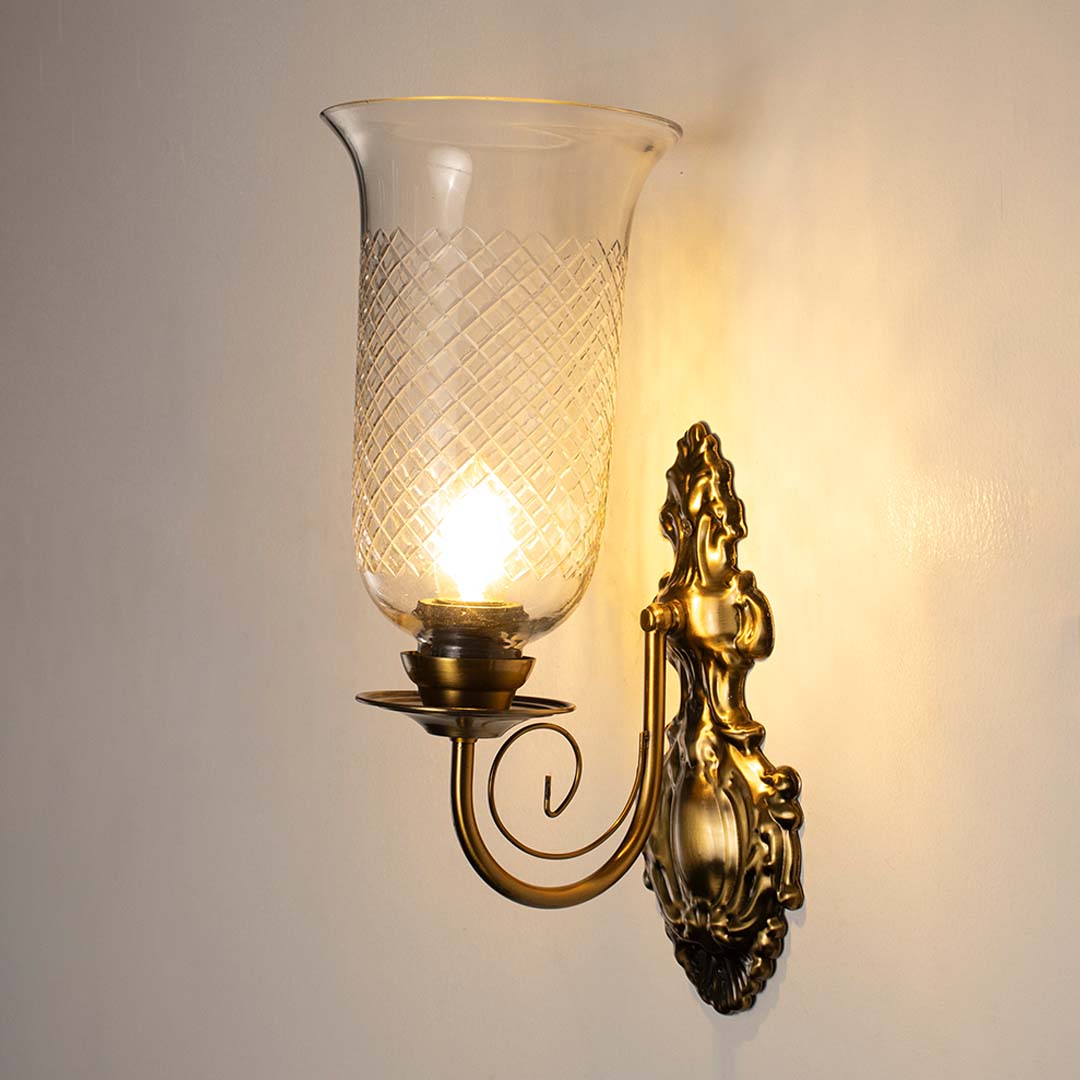 Illuminated antique brass wall sconce with tall glass shade turned on glowing against a clean white background.