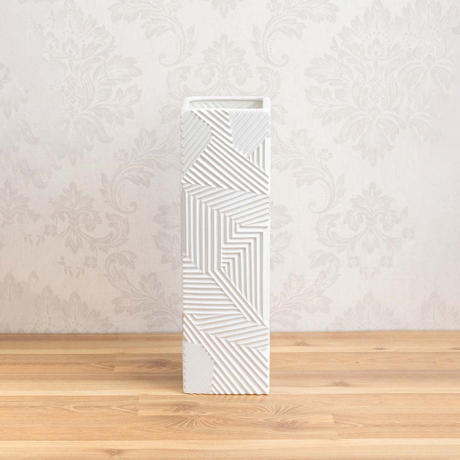 Tall white ceramic square pillar vase featuring relief geometric linear patterns displayed on a natural wood tabletop.