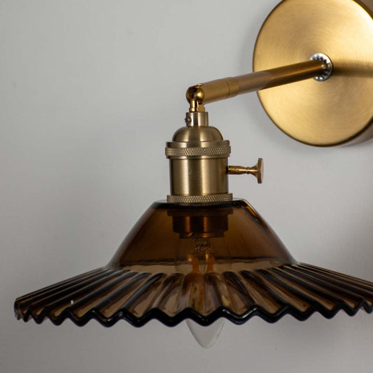 Decorative pleated dark glass wall sconce with brass metal base fixed against soft wall background perfect for luxury interior lighting setup.
