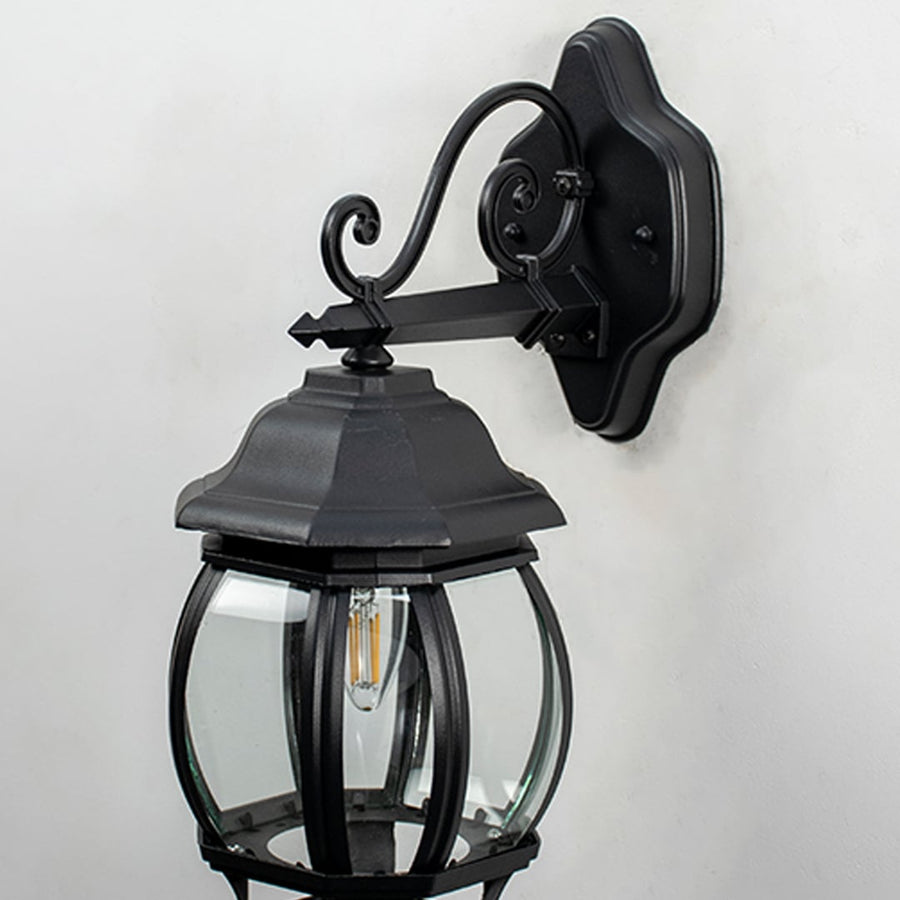 Closeup view of outdoor wall lantern showing detailed black metal arm mounting plate and clear glass panels with visible bulb holder and textured finish.