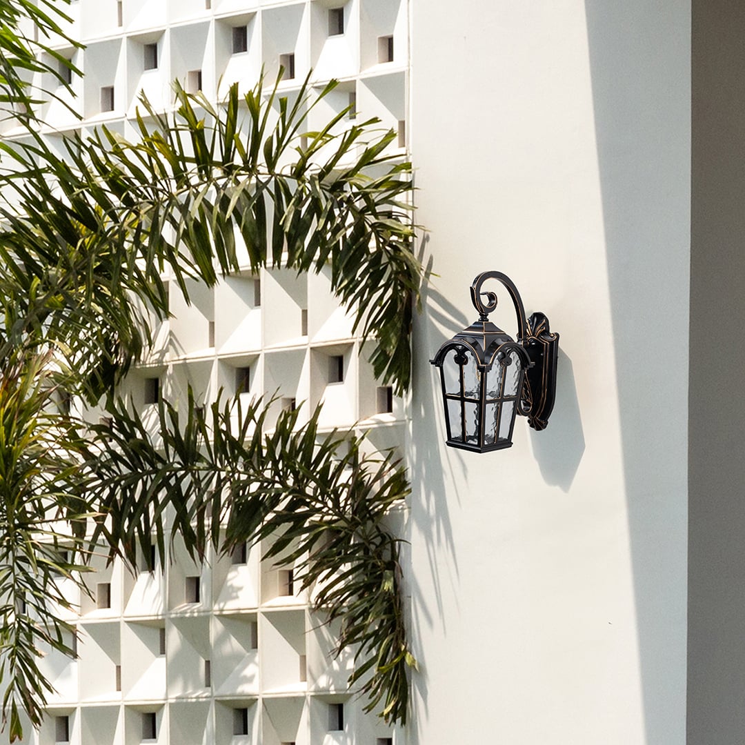 Black metal outdoor wall light lifestyle image with textured glass shade mounted on white villa wall with palm trees.