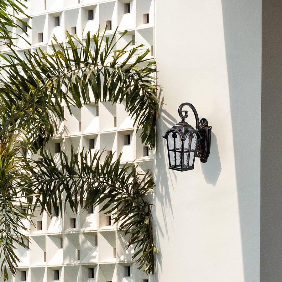 Black metal outdoor wall light lifestyle image with textured glass shade mounted on white villa wall with palm trees.