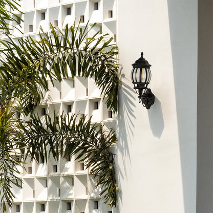 Black metal traditional wall lamp with vertical ribbed glass shade installed on white decorative breeze block wall near palm trees.