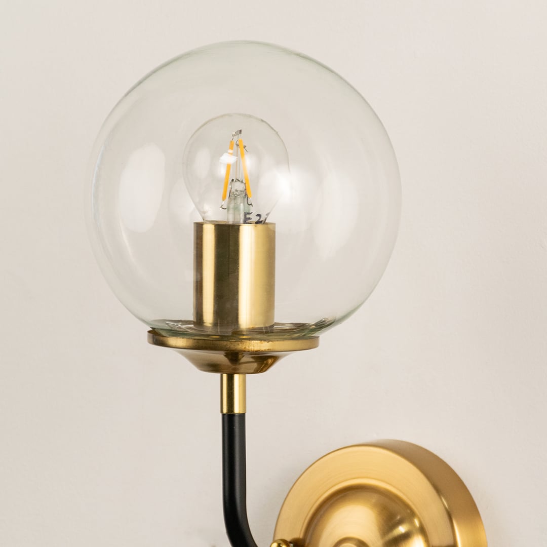 A metal wall light with a clear round glass globe and gold mount glowing on a beige surface creating warm ambient lighting and adding stylish contemporary décor.