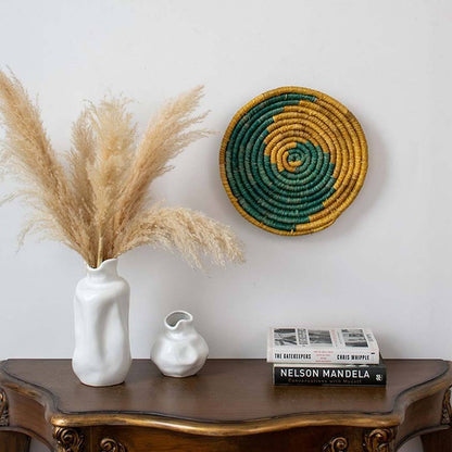 Decorative setup with pampas grass in a white vase, smaller white vase, books, and a woven wall art piece on a wooden surface.