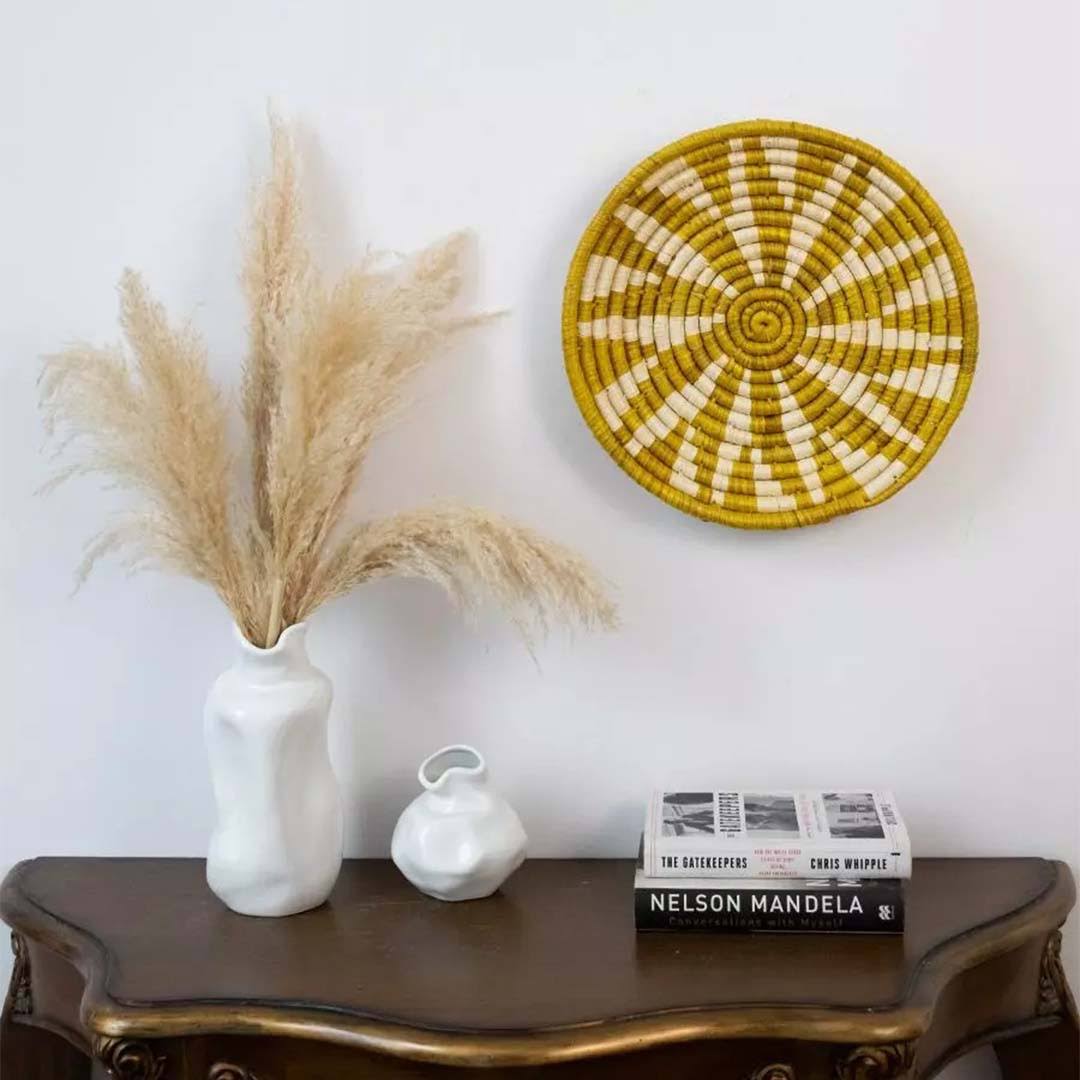 Decorative setup with a vase of pampas grass, books, and a woven wall hanging on a wooden console table.