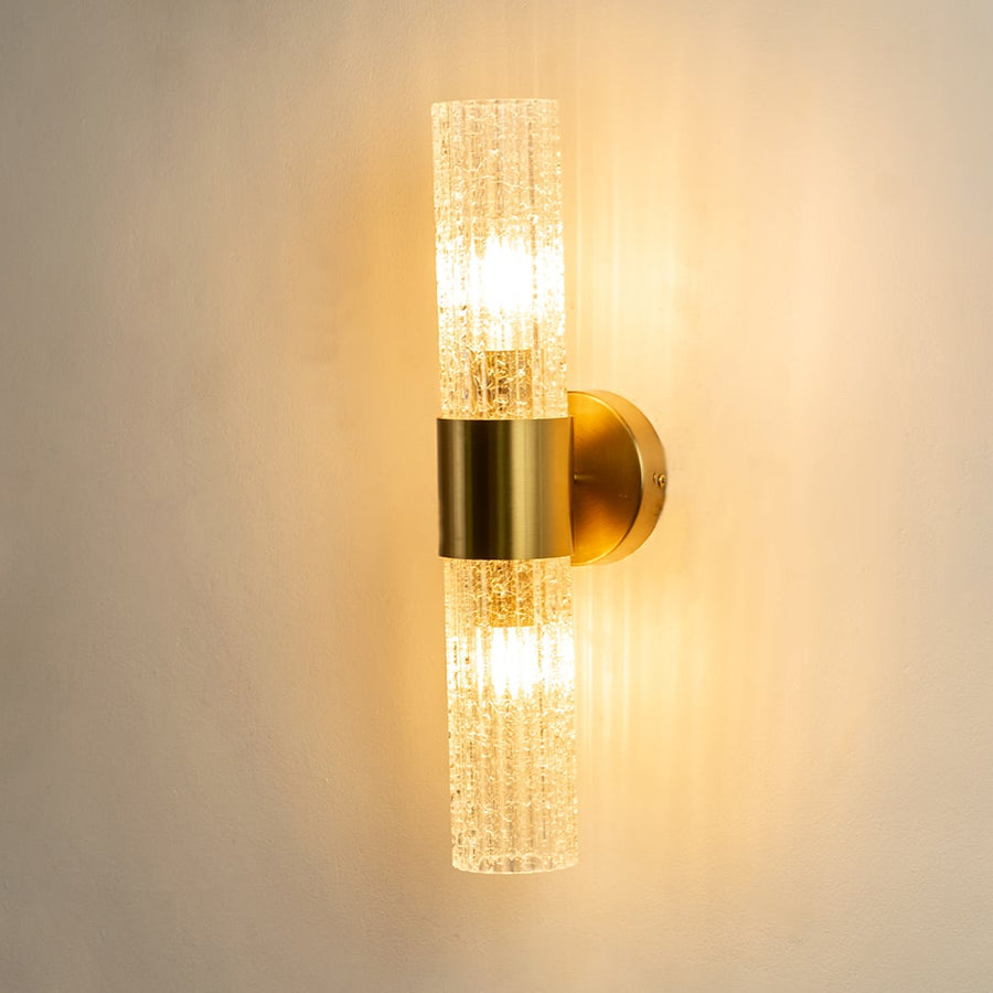A gold wall sconce with two textured cylindrical glass shades shown with lights on against a white background highlighting its warm ambient glow and vertical design.