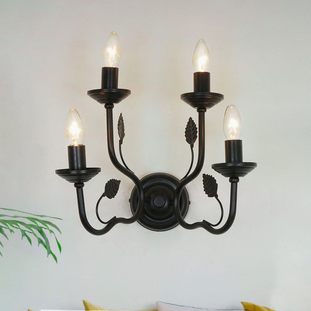A classic black four light wall sconce glowing warmly in a soft neutral living space adding elegant lighting with curved metal arms and leaf accents.