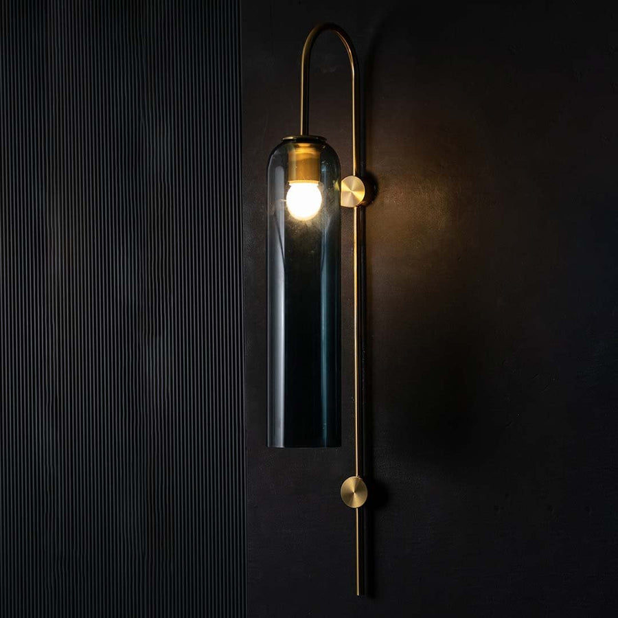 Gold and teal glass wall light glowing warmly on a dark background showcasing its long cylindrical shade and soft ambient modern lighting effect.