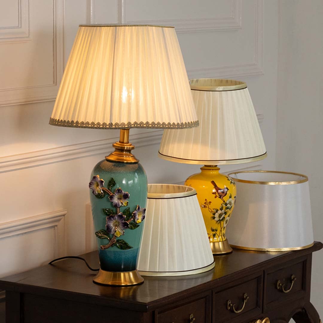Beige pleated shade floral ceramic table lamp on wooden sideboard with light on for vintage decorative décor.